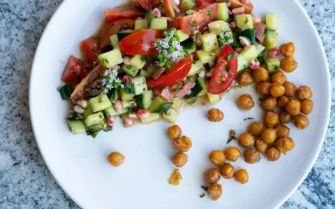 Salade colorée de tomates, concombres et pois chiches, garnie de petites fleurs blanches. Des pois chiches rôtis sont disposés à côté.