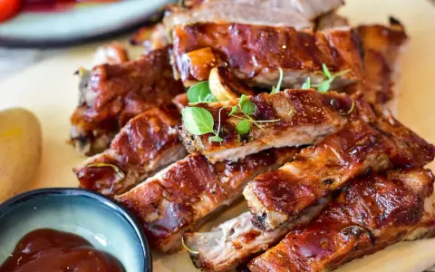 Délicieux ribs au four, nappés d'une sauce barbecue brillante, servis avec une petite coupelle de sauce et des herbes fraîches.