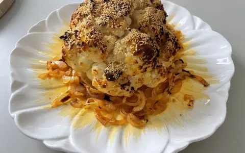 Un chou-fleur entier, rôti au four, est servi sur une assiette blanche ornée d'oignons caramélisés. Le chou-fleur est saupoudré de graines de sésame et d'épices.