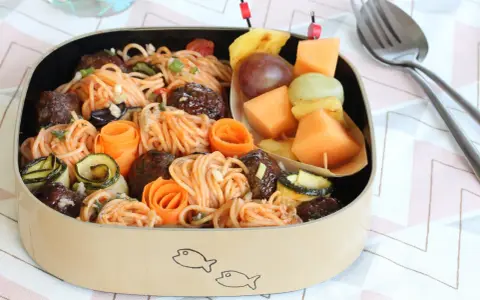 Bento box avec spaghetti et boulettes, garni de courgettes et carottes roulées. Une brochette de fruits complète cette idée repas à emporter au travail.