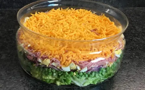 Salade à étage colorée dans un bol en verre : laitue, petits pois, oignons rouges, jambon, œufs durs et fromage râpé.