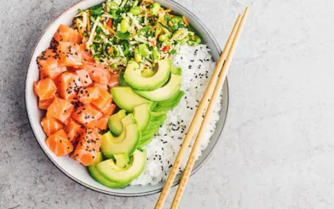 Un délicieux base poke bowl avec saumon cru, avocat, riz blanc et légumes croquants, prêt à être dégusté avec des baguettes.