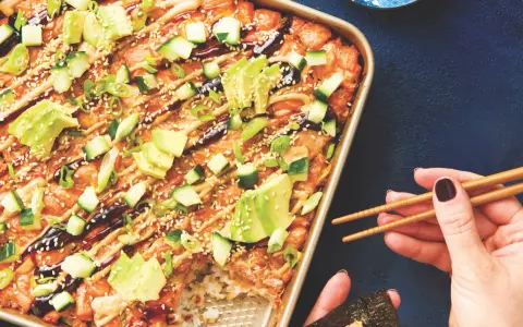 Un délicieux sushi bake dans un plat doré, garni d'avocat, concombre, oignons verts et graines de sésame. Une main tient des baguettes pour servir.