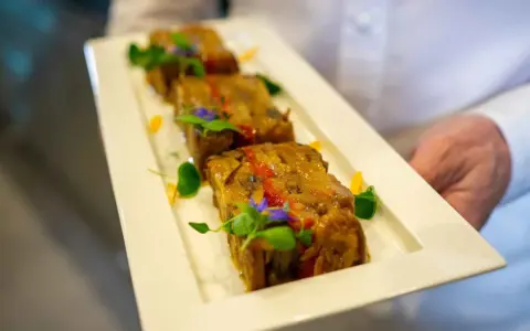 Trois tranches de terrine de légumes, garnies de fleurs comestibles et de verdure, présentées sur une assiette blanche par un serveur.