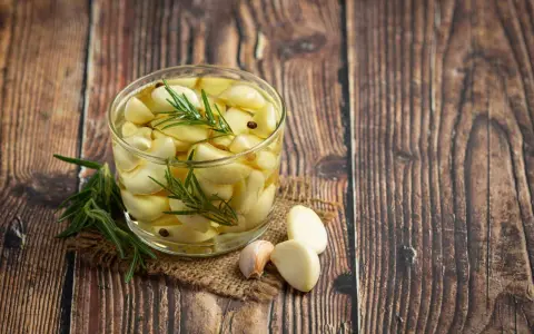 Un bocal de **ail confit** avec des brins de romarin et quelques grains de poivre, posé sur une toile de jute, sur un fond de bois rustique.