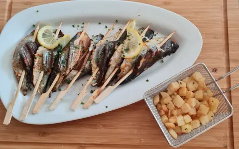 Sardines grillées sur brochettes, garnies de citron et persil, accompagnées de pommes de terre sautées dans un panier métallique.