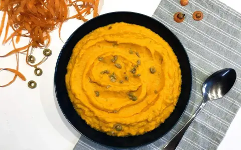 Un bol de purée de carotte onctueuse, garnie de graines, à côté d'une cuillère et de carottes râpées.