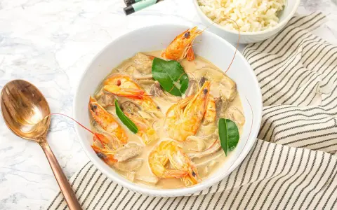 Un plat de sauté crevettes thaï lait de coco, garni de feuilles vertes, servi avec du riz et une cuillère en cuivre.