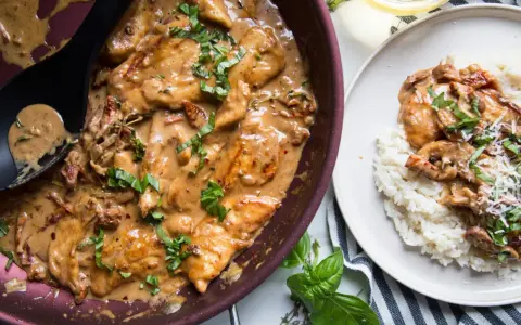 Poulet en sauce tomate séchée crème, garni de basilic frais, servi dans une poêle et sur une assiette de riz.