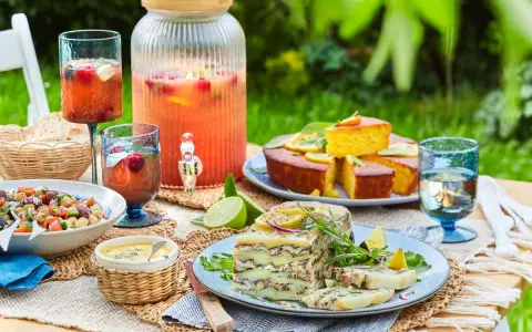 Un délicieux repas d'été entre amis : salade, terrine, gâteau, et une grande carafe de boisson fruitée, le tout sur une table dressée en extérieur.