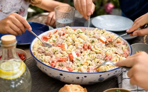 Un grand plat de salade de pâtes au surimi, tomates et thon, servi à l'extérieur. Des mains servent la salade.