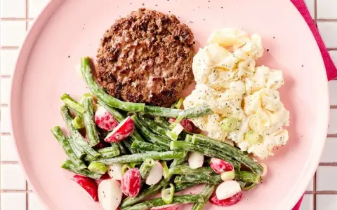 Un hamburger savoureux avec une salade de haricots verts et radis, et une salade de pommes de terre crémeuse sur une assiette rose.