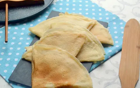 Pile de crêpes dorées, prêtes à être garnies. La pâte à crêpe vegan est légère et aérienne. Un ustensile en bois est à côté.