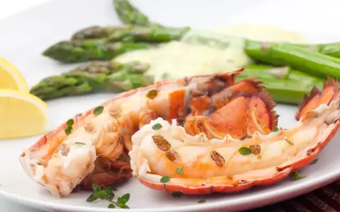 Queue de homard grillée, servie avec des asperges et une sauce homard crémeuse, garnie de quartiers de citron et de thym frais.