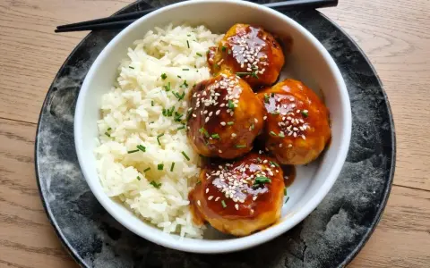 Délicieuses boulettes de poulet asiatique nappées d'une sauce sucrée et salée, servies avec du riz blanc et parsemées de graines de sésame.