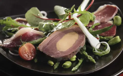 Une délicieuse salade landaise avec du foie gras, du magret de canard, des asperges et des tomates cerises sur une assiette noire.