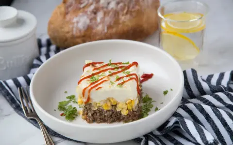 Un délicieux pâté chinois servi dans une assiette blanche, garni de persil et de ketchup. Un verre d'eau citronnée et du pain complètent ce plat réconfortant.