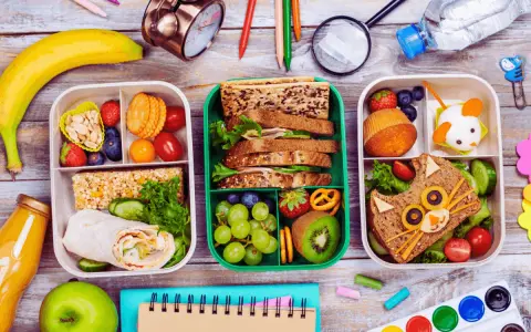 Trois boîtes repas à emporter faites maison, remplies de fruits, sandwichs créatifs et barres de céréales. Parfait pour un pique-nique ou un déjeuner sur le pouce.