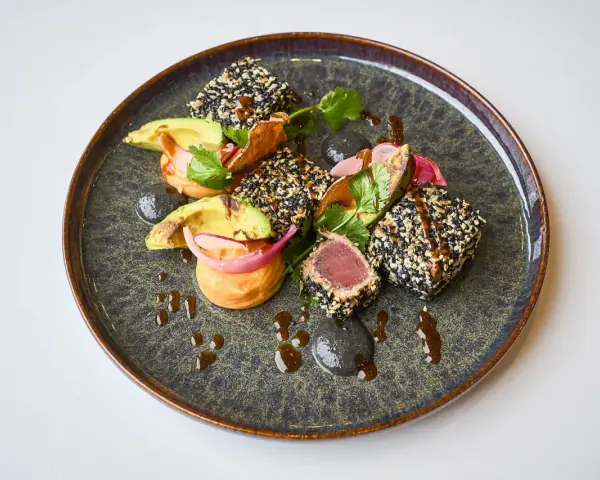 Un délicieux tataki de thon enrobé de sésame, servi avec de l'avocat, des oignons rouges et une sauce onctueuse sur une assiette texturée.