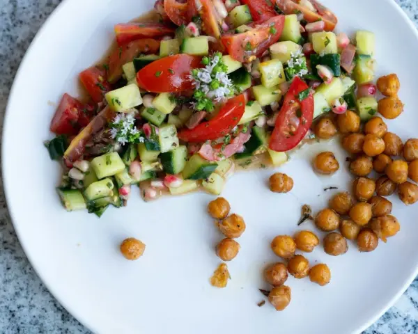 Salade colorée de tomates, concombres et pois chiches, garnie de petites fleurs blanches. Des pois chiches rôtis sont disposés à côté.