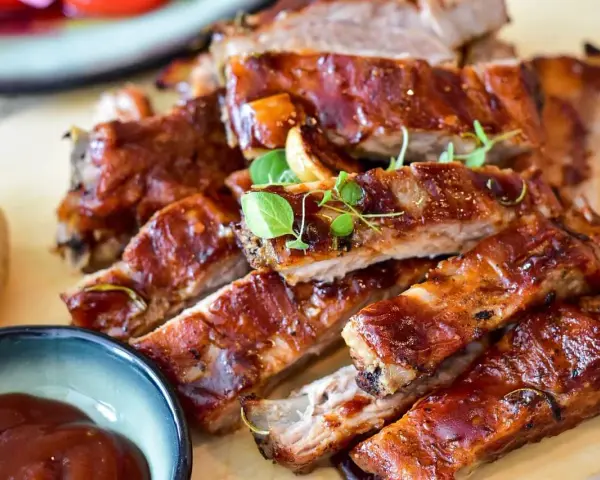 Délicieux ribs au four, nappés d'une sauce barbecue brillante, servis avec une petite coupelle de sauce et des herbes fraîches.