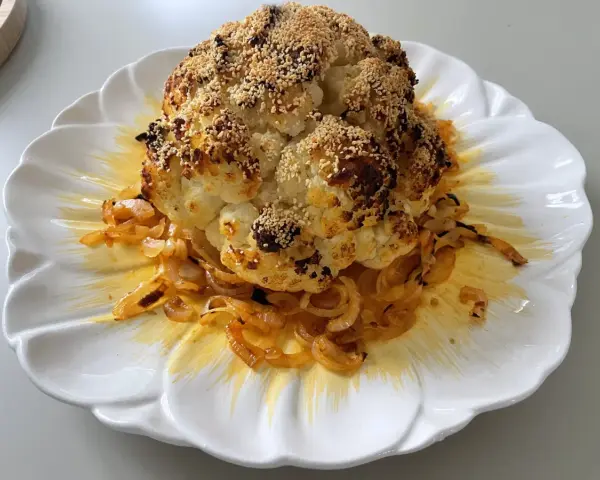 Un chou-fleur entier, rôti au four, est servi sur une assiette blanche ornée d'oignons caramélisés. Le chou-fleur est saupoudré de graines de sésame et d'épices.