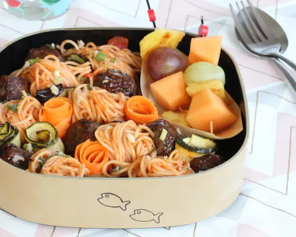 Bento box avec spaghetti et boulettes, garni de courgettes et carottes roulées. Une brochette de fruits complète cette idée repas à emporter au travail.