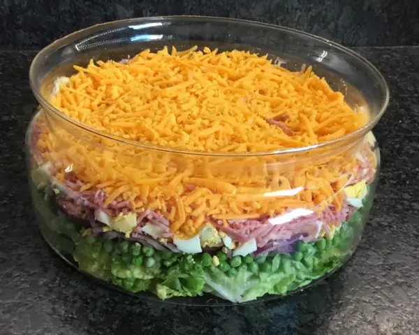Salade à étage colorée dans un bol en verre : laitue, petits pois, oignons rouges, jambon, œufs durs et fromage râpé.