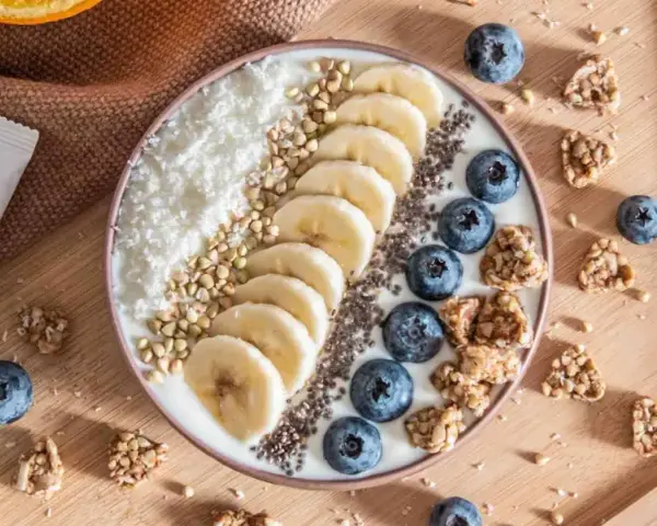 Un bol de smoothie garni de bananes, myrtilles, noix de coco râpée et graines. Une recette pas cher et saine pour un petit-déjeuner gourmand.
