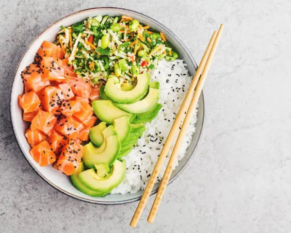 Un délicieux poke bowl santé avec saumon cru, avocat, riz blanc et légumes croquants, prêt à être dégusté avec des baguettes.