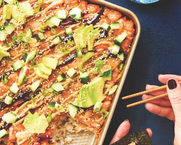 Un délicieux sushi bake dans un plat doré, garni d'avocat, concombre, oignons verts et graines de sésame. Une main tient des baguettes pour servir.