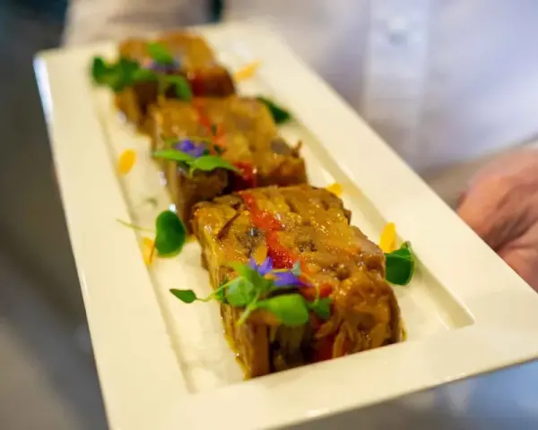Trois tranches de terrine de légumes, garnies de fleurs comestibles et de verdure, présentées sur une assiette blanche par un serveur.