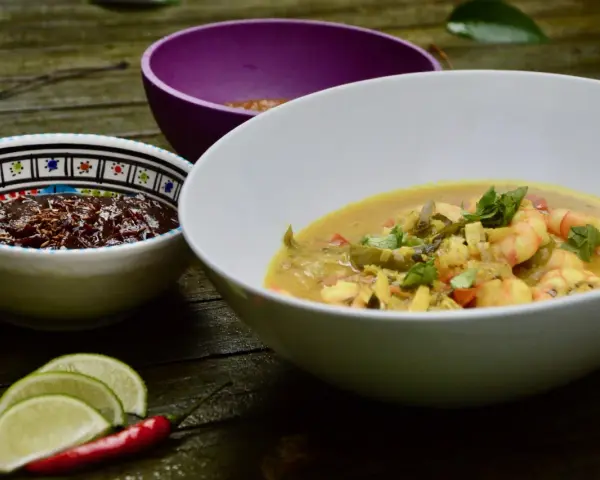 Un plat de crevette curry coco crémeux, garni de coriandre fraîche, servi avec des quartiers de citron vert et un piment rouge.