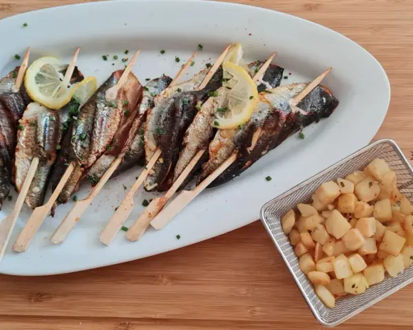 Sardines grillées sur brochettes, garnies de citron et persil, accompagnées de pommes de terre sautées dans un panier métallique.