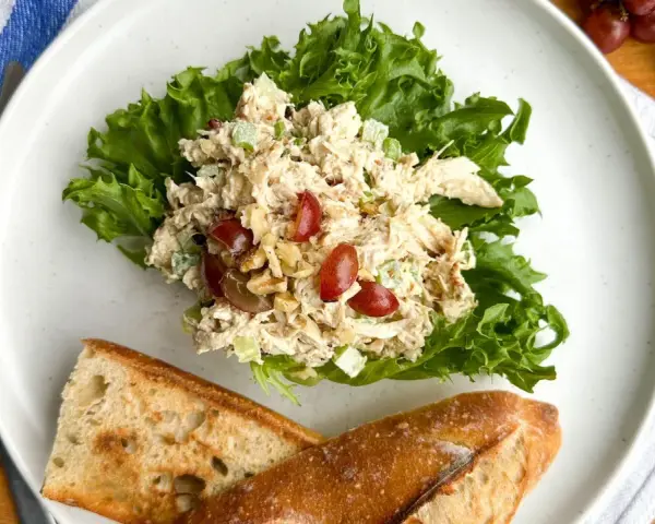 Salade de poulet froid mayonnaise sur laitue, avec des raisins et des noix, servie avec du pain croustillant.