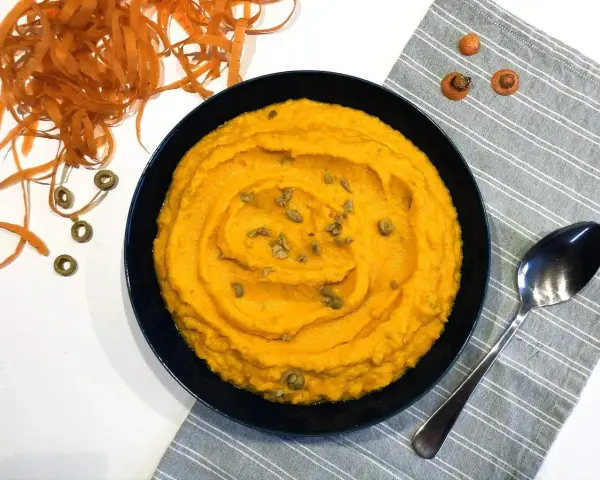 Un bol de purée de carotte onctueuse, garnie de graines, à côté d'une cuillère et de carottes râpées.