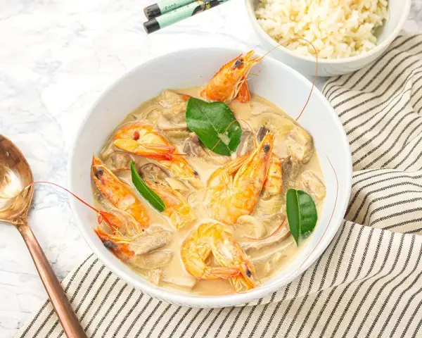 Un plat de sauté crevettes thaï lait de coco, garni de feuilles vertes, servi avec du riz et une cuillère en cuivre.