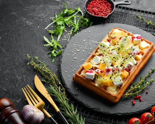 Gaufre salée garnie de fromage, de dés de mangue et de pousses vertes, sur une ardoise noire.