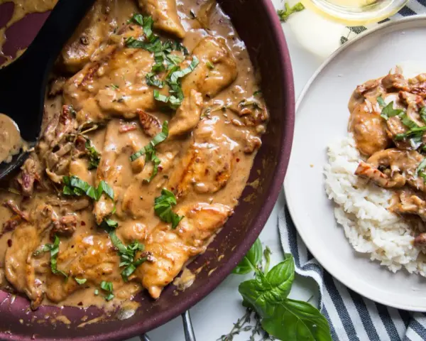 Poulet en sauce tomate séchée crème, garni de basilic frais, servi dans une poêle et sur une assiette de riz.