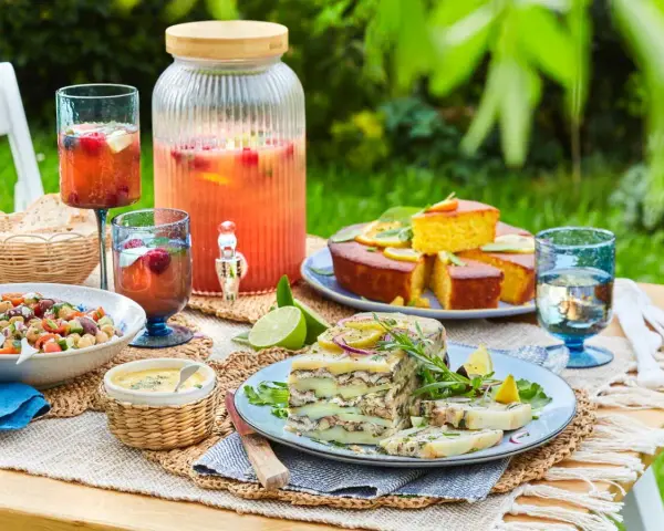 Un délicieux repas d'été entre amis : salade, terrine, gâteau, et une grande carafe de boisson fruitée, le tout sur une table dressée en extérieur.