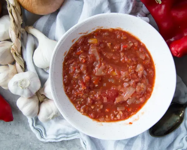 Un bol de salsa facile, avec des tomates, des oignons et des poivrons rouges. Des gousses d'ail et un oignon sont disposés autour.