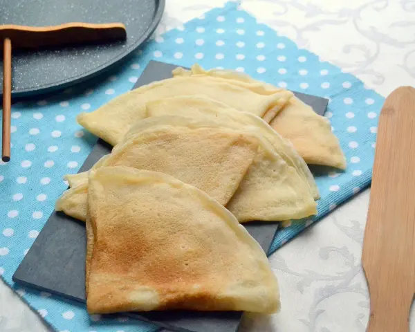 Pile de crêpes dorées, prêtes à être garnies. La pâte à crêpe vegan est légère et aérienne. Un ustensile en bois est à côté.