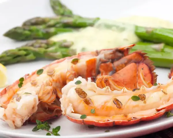 Queue de homard grillée, servie avec des asperges et une sauce homard crémeuse, garnie de quartiers de citron et de thym frais.