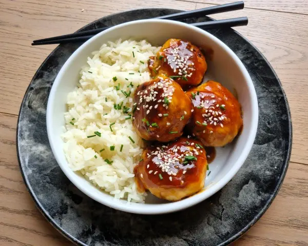 Délicieuses boulettes de poulet asiatique nappées d'une sauce sucrée et salée, servies avec du riz blanc et parsemées de graines de sésame.
