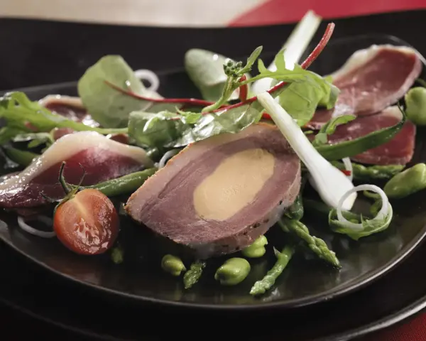 Une délicieuse salade landaise avec du foie gras, du magret de canard, des asperges et des tomates cerises sur une assiette noire.