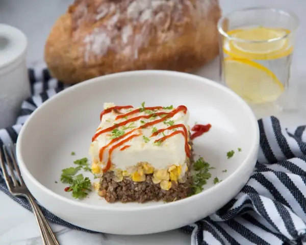 Un délicieux pâté chinois servi dans une assiette blanche, garni de persil et de ketchup. Un verre d'eau citronnée et du pain complètent ce plat réconfortant.