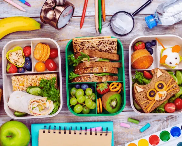 Trois boîtes repas à emporter faites maison, remplies de fruits, sandwichs créatifs et barres de céréales. Parfait pour un pique-nique ou un déjeuner sur le pouce.