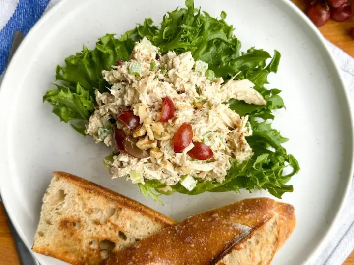 Salade de poulet froid mayonnaise sur laitue, avec des raisins et des noix, servie avec du pain croustillant.