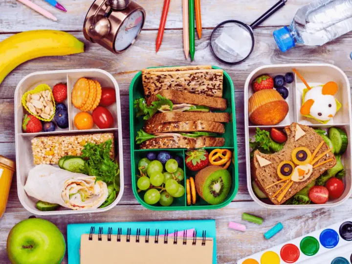 Trois boîtes repas à emporter faites maison, remplies de fruits, sandwichs créatifs et barres de céréales. Parfait pour un pique-nique ou un déjeuner sur le pouce.