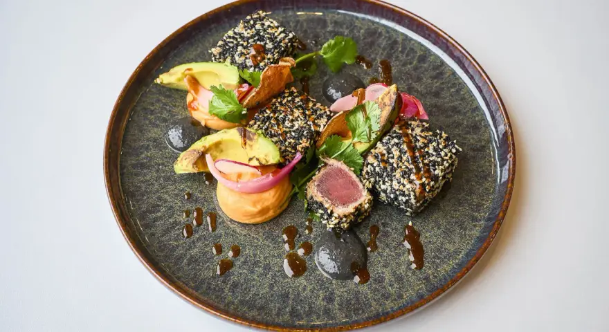 Un délicieux tataki de thon enrobé de sésame, servi avec de l'avocat, des oignons rouges et une sauce onctueuse sur une assiette texturée.